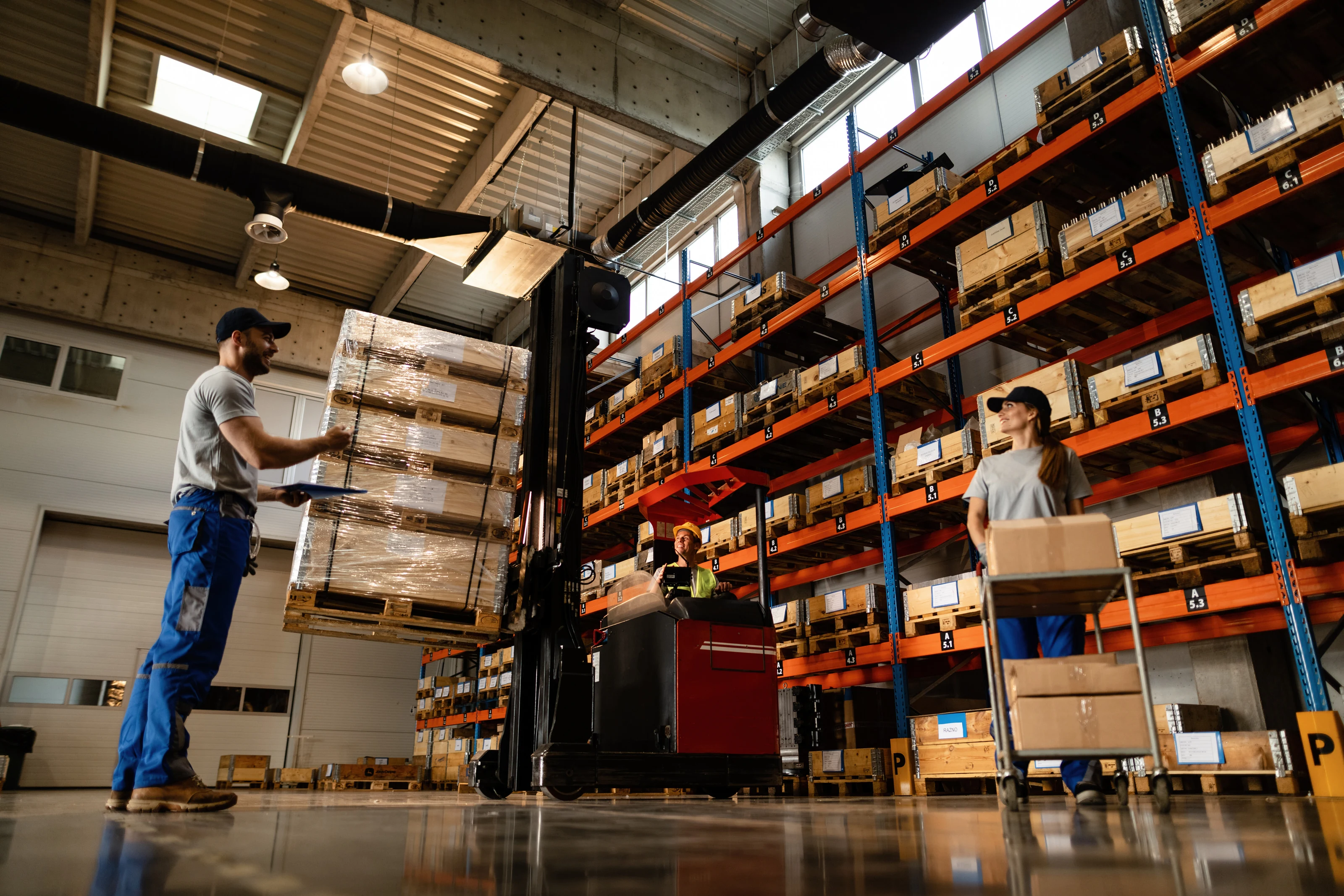 Warehouse staff moving pallets of stock between tall storage racks.