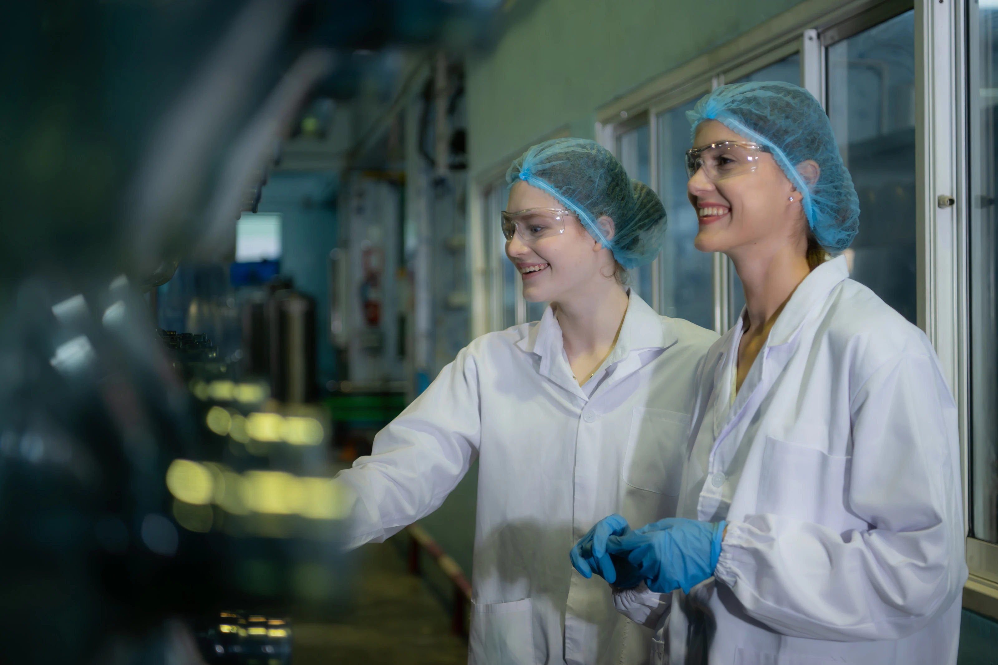 Two technicians in white coats and hairnets inspecting equipment on a pharmaceutical production line.