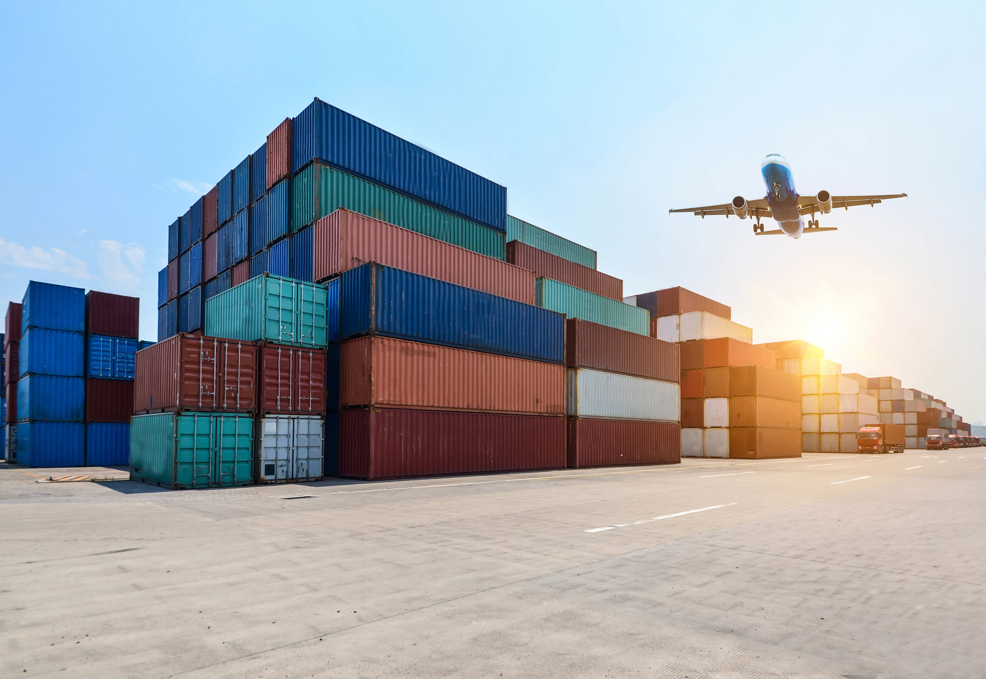 Stacked shipping containers at a port with an aircraft climbing overhead at sunset.