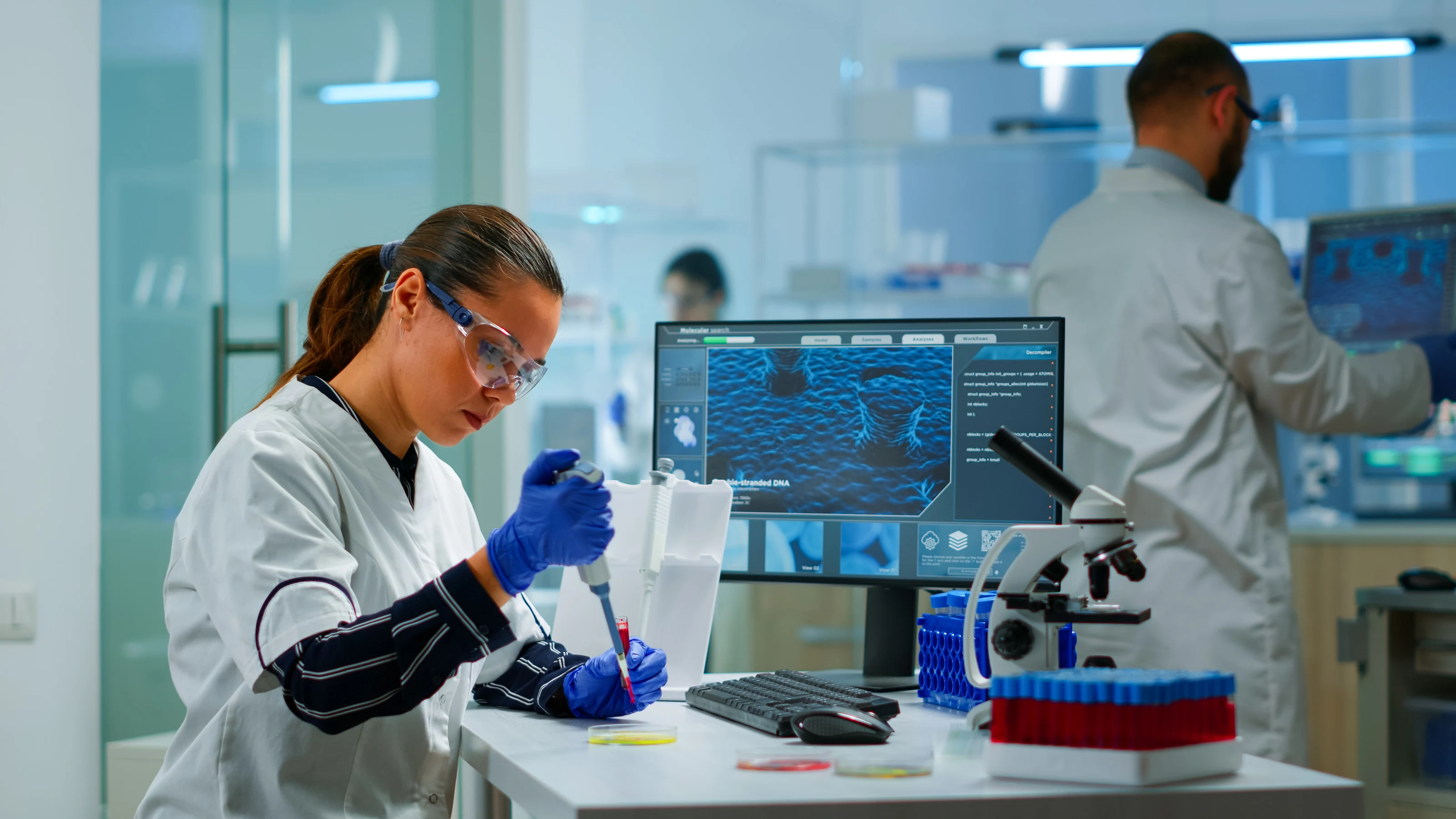 A scientist in safety glasses pipetting a sample at a laboratory bench, with a colleague working behind.