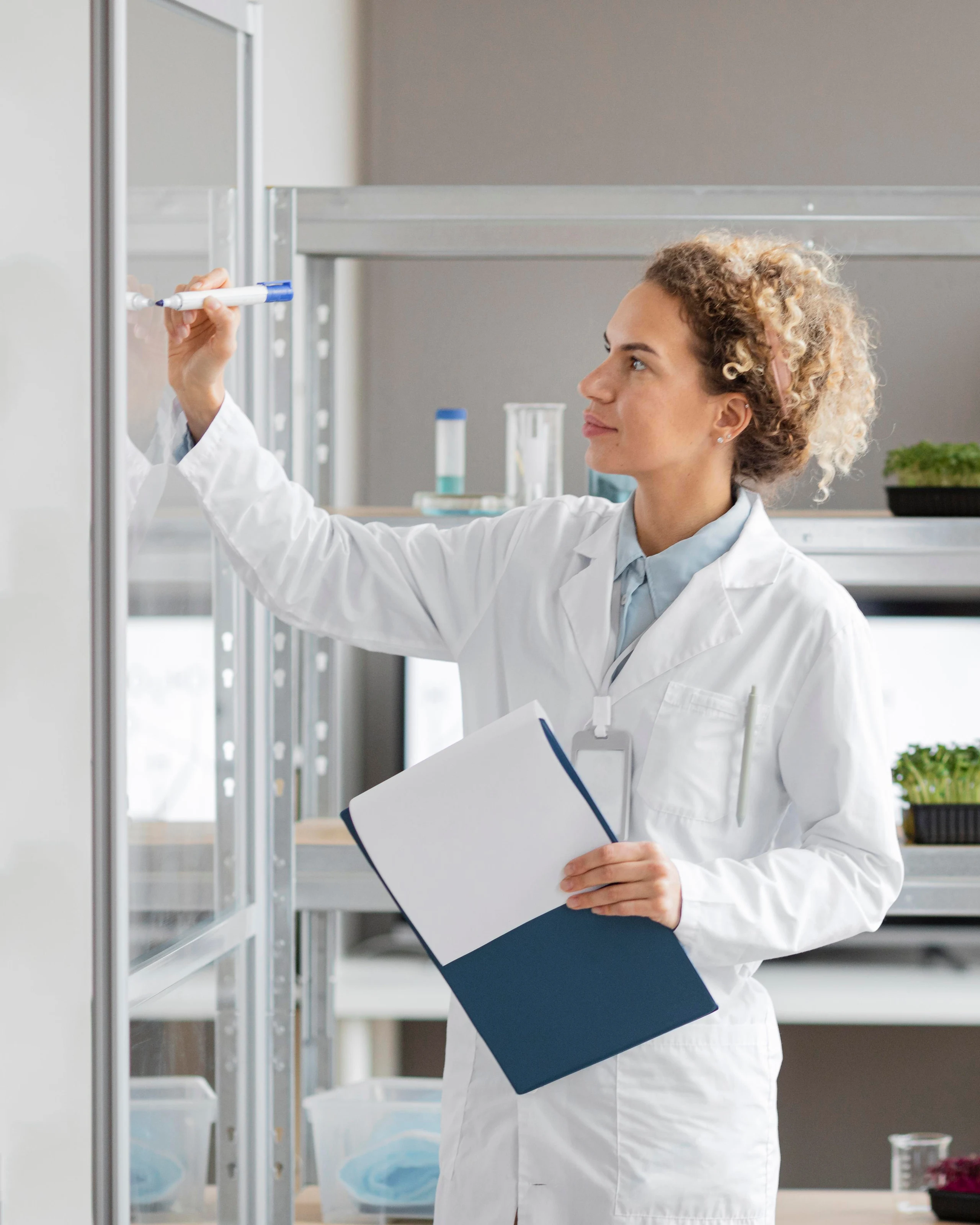A researcher in a white coat writing up results beside laboratory shelving.