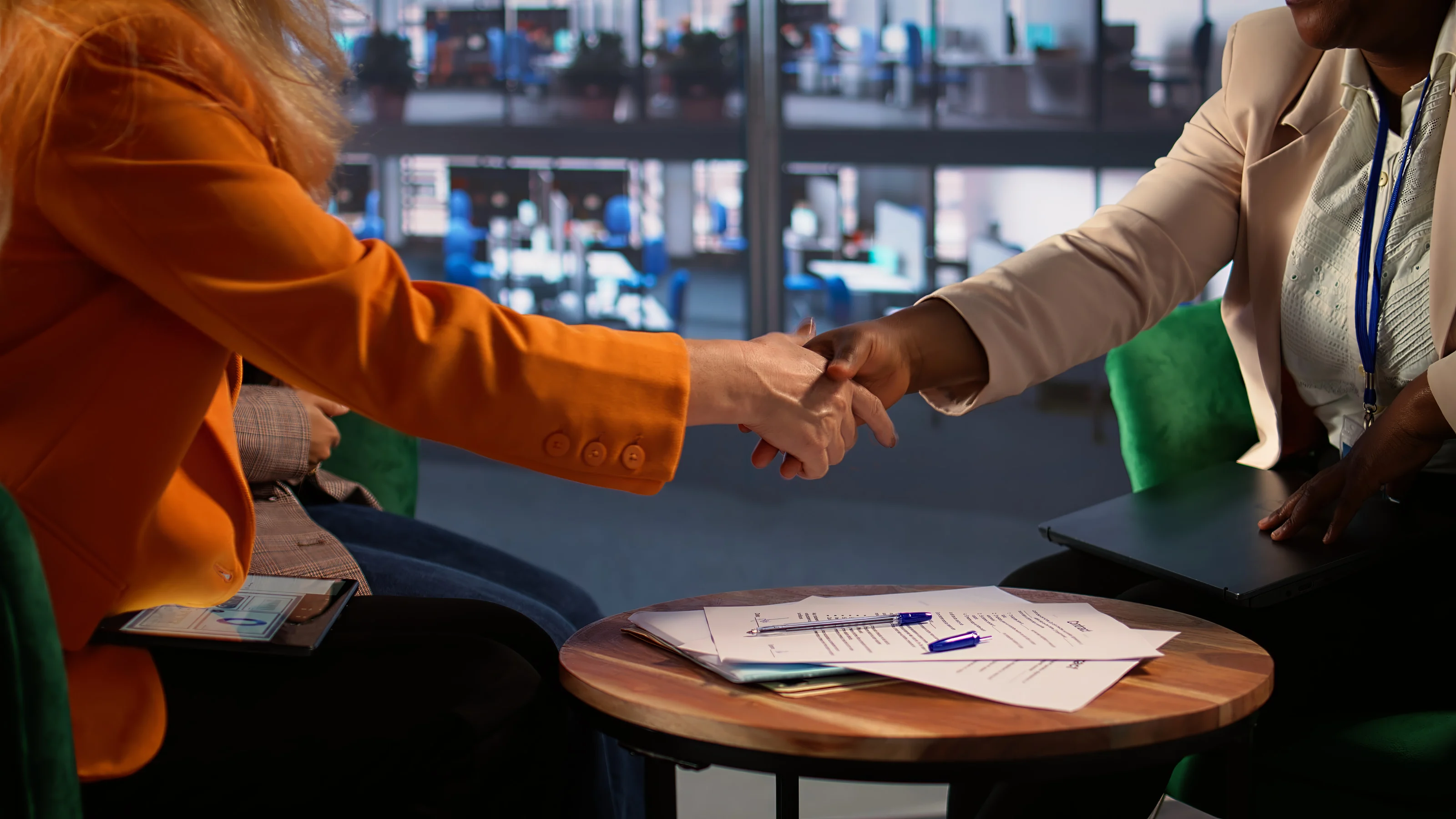Two people shaking hands across a table with signed papers on it.