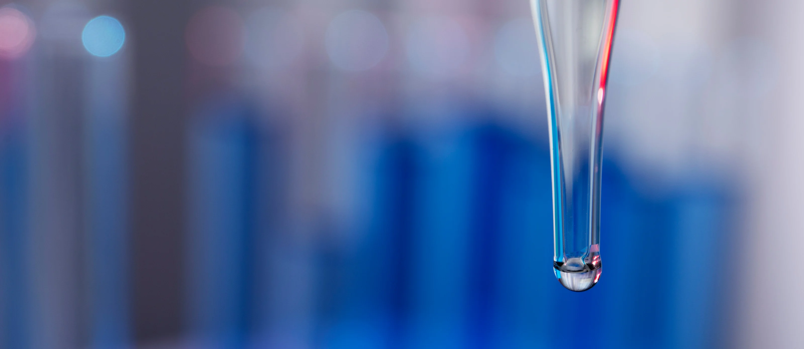 Close view of a glass pipette tip holding a single droplet, against blurred blue laboratory glassware.