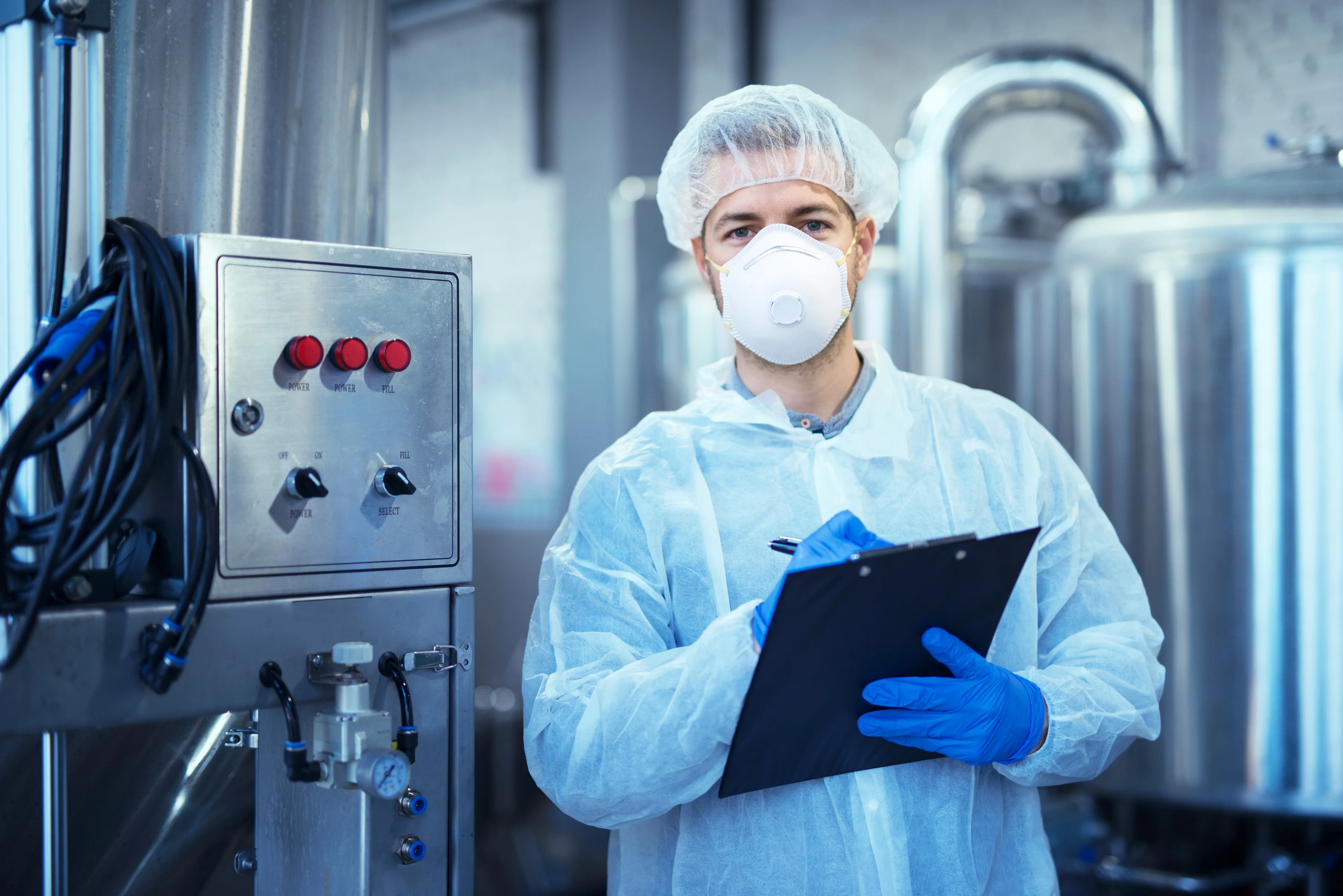 A technician in a white protective uniform, hairnet and face mask standing at an industrial production machine.