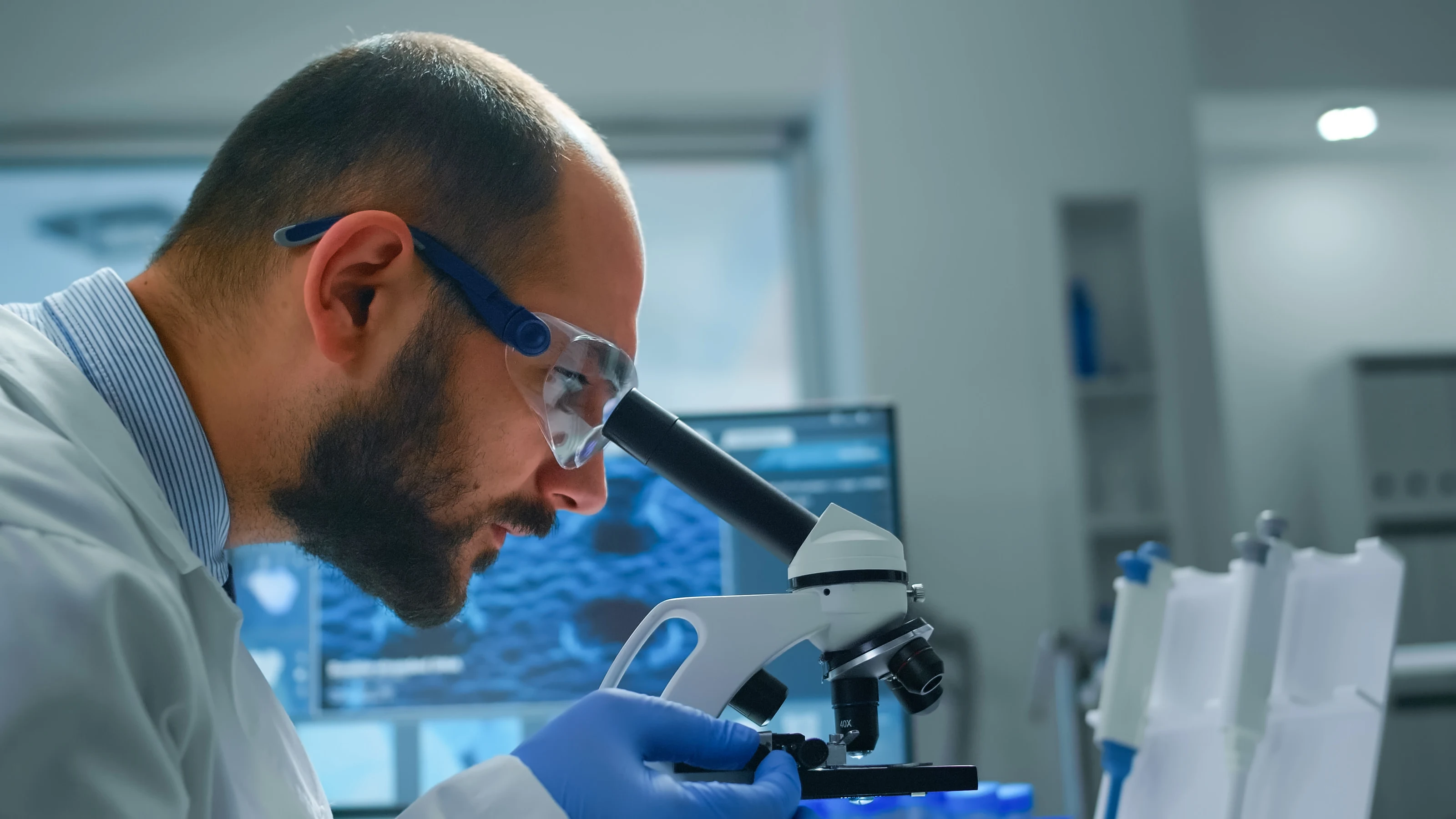A research scientist in safety glasses examining a sample under a microscope.