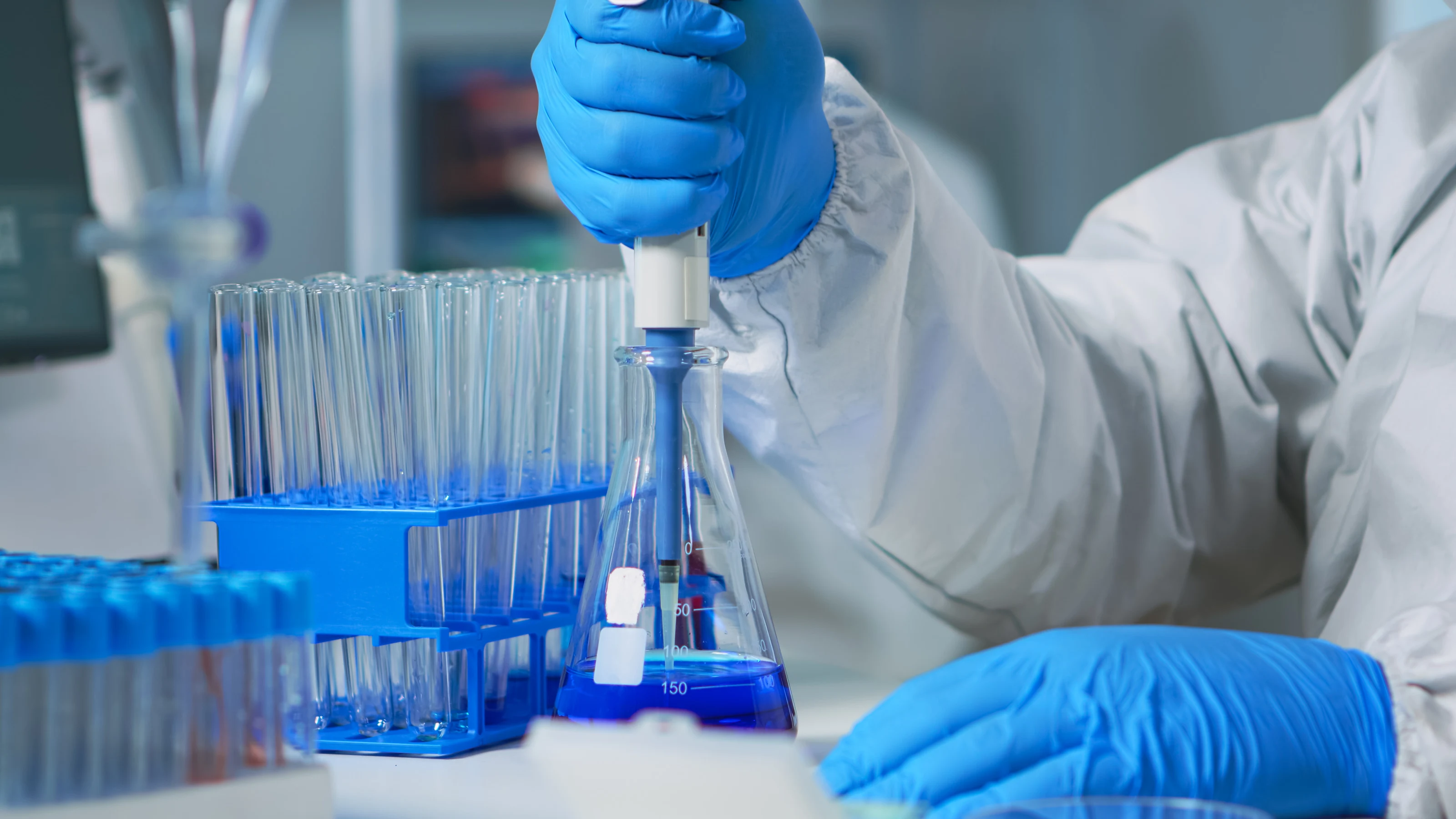 Gloved hands using a micropipette over a flask beside a rack of sample tubes.