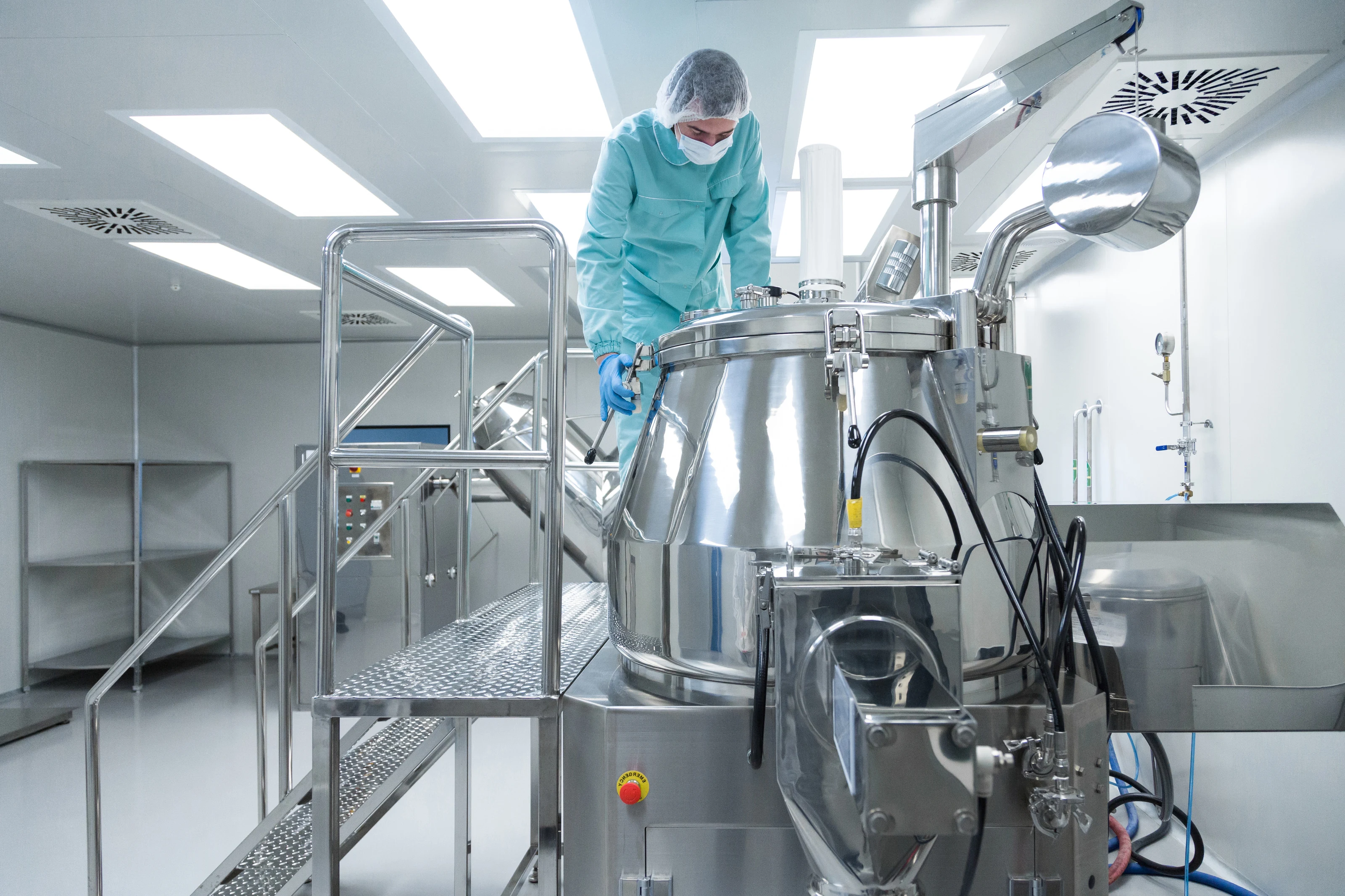 An operator in protective clothing working at a stainless steel vessel in a sterile pharmaceutical production room.