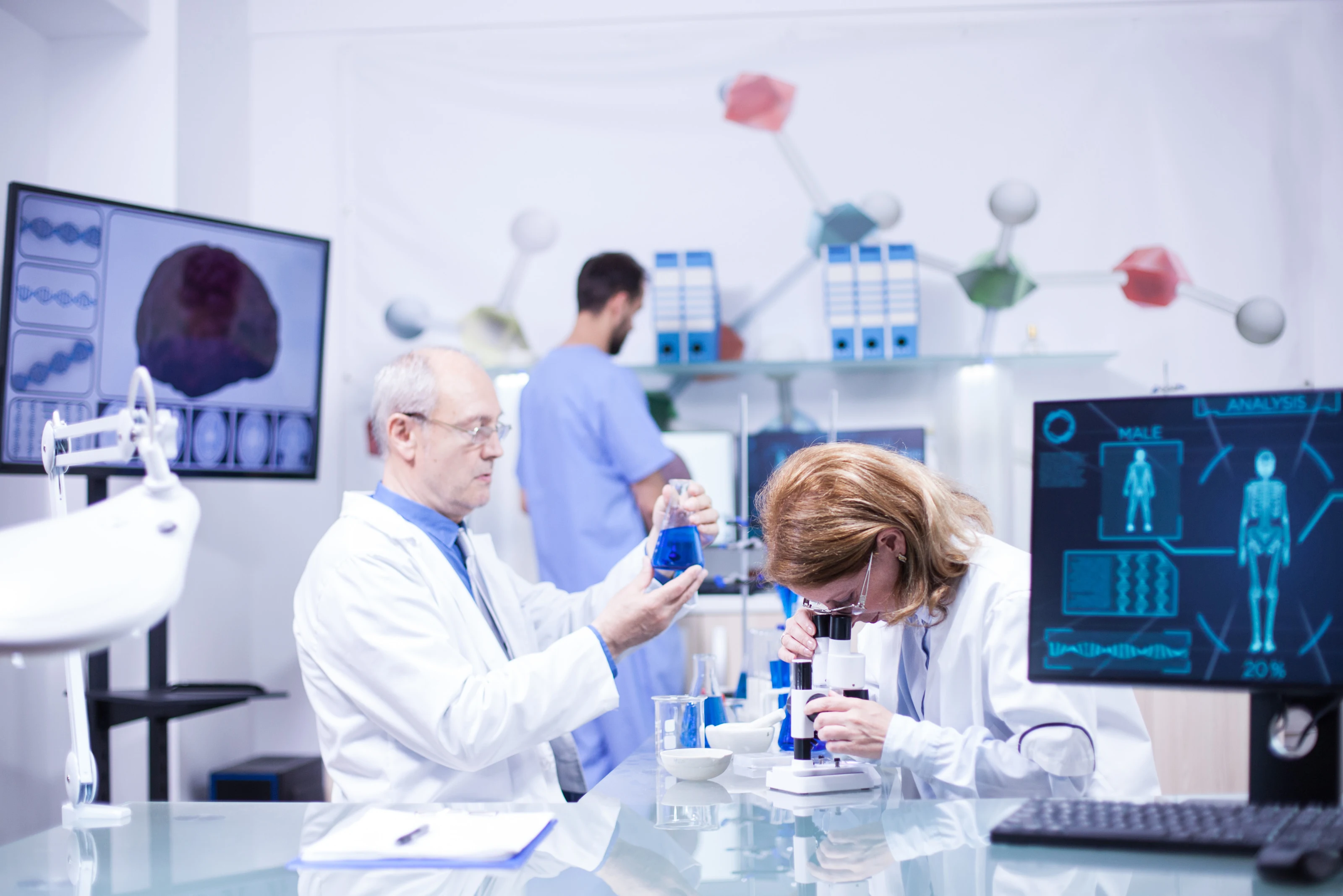Two researchers working at a microscope in a laboratory, with a molecular model behind them.