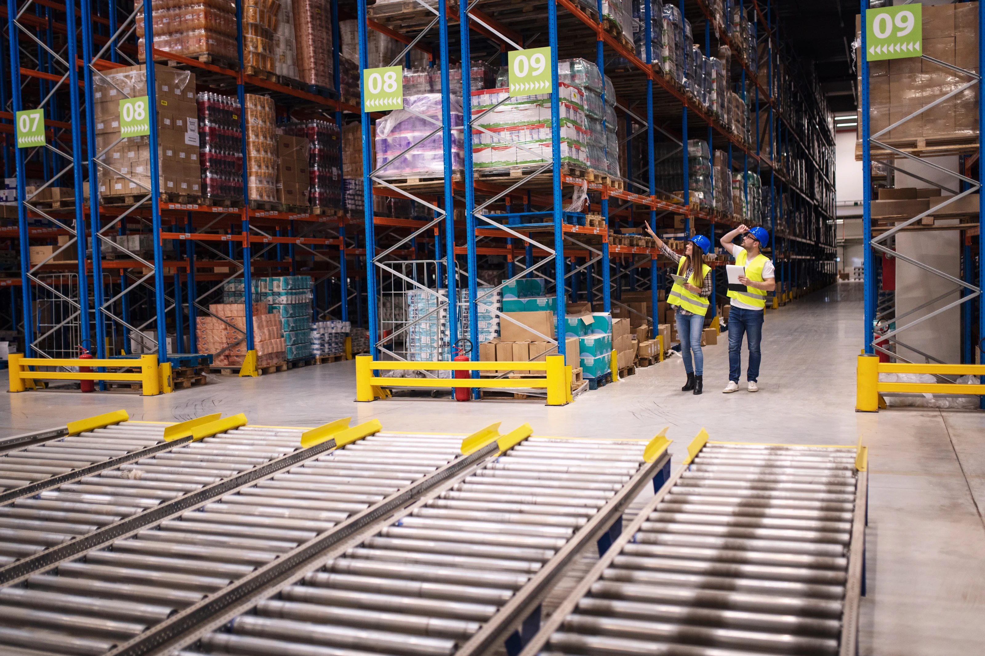 Two workers in high-visibility jackets walking an aisle of a distribution warehouse.