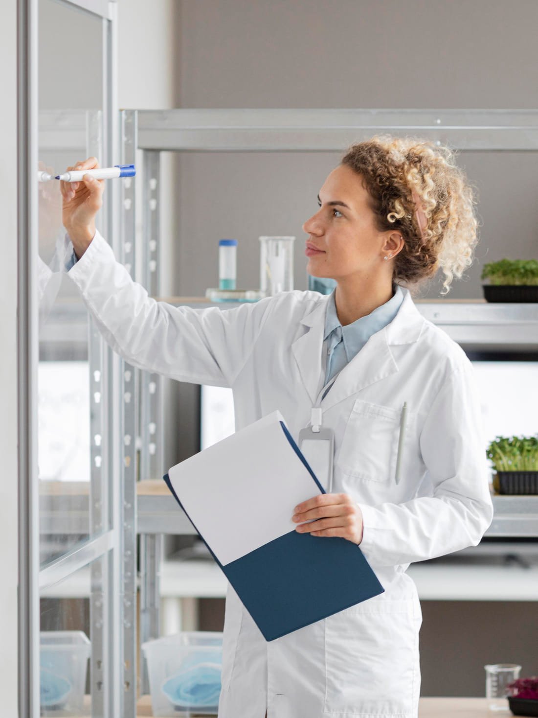A researcher in a white coat writing up results beside laboratory shelving.